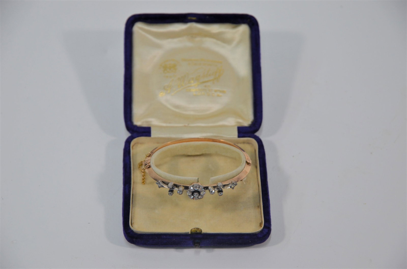 An early 20th century diamond set gold bangle: An early 20th century yellow gold bangle with diamond set central daisy cluster and diamond set tendrils, half hinged, safety chain attached, 6 cm diam, approx 10g all in, in original fitted blue velv