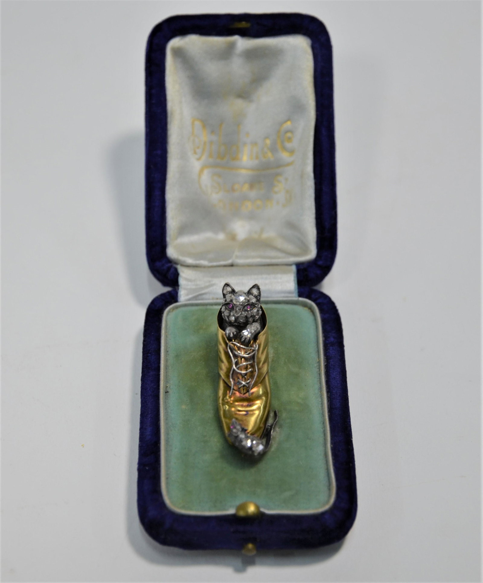A gold and diamond set antique brooch in the form of a: An unusual antique gold and diamond set brooch in the form of a well-modelled and detailed boot with untied silver laces, with a diamond set cat with cabochon ruby eyes peeping out of the top of the b