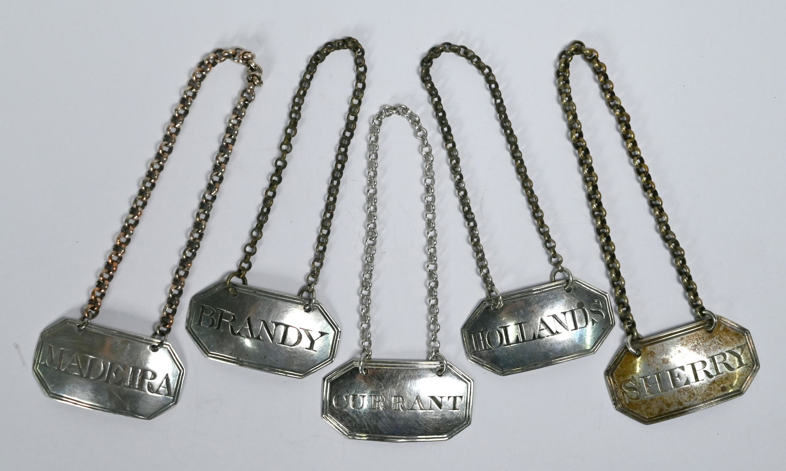 Five George III silver decanter labels: Four George III silver decanter labels of near-matching design, for Holland, Madeira, Sherry and Brandy, Edward Mayfield, London 1800, to/w a similar label for Currant, Thomas Johnson, London 1807 (5)