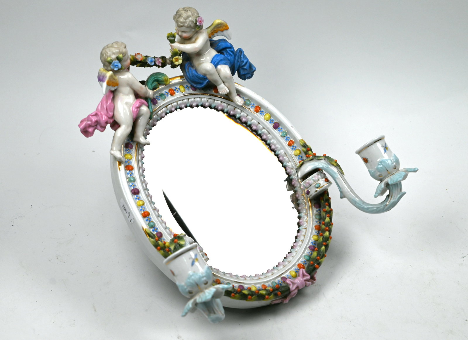A Dresden porcelain oval girandole mirror: A Dresden porcelain oval girandole mirror with twin candle-sconces, surmounted by cherubs with floral garland, 29 x 28 cm overall