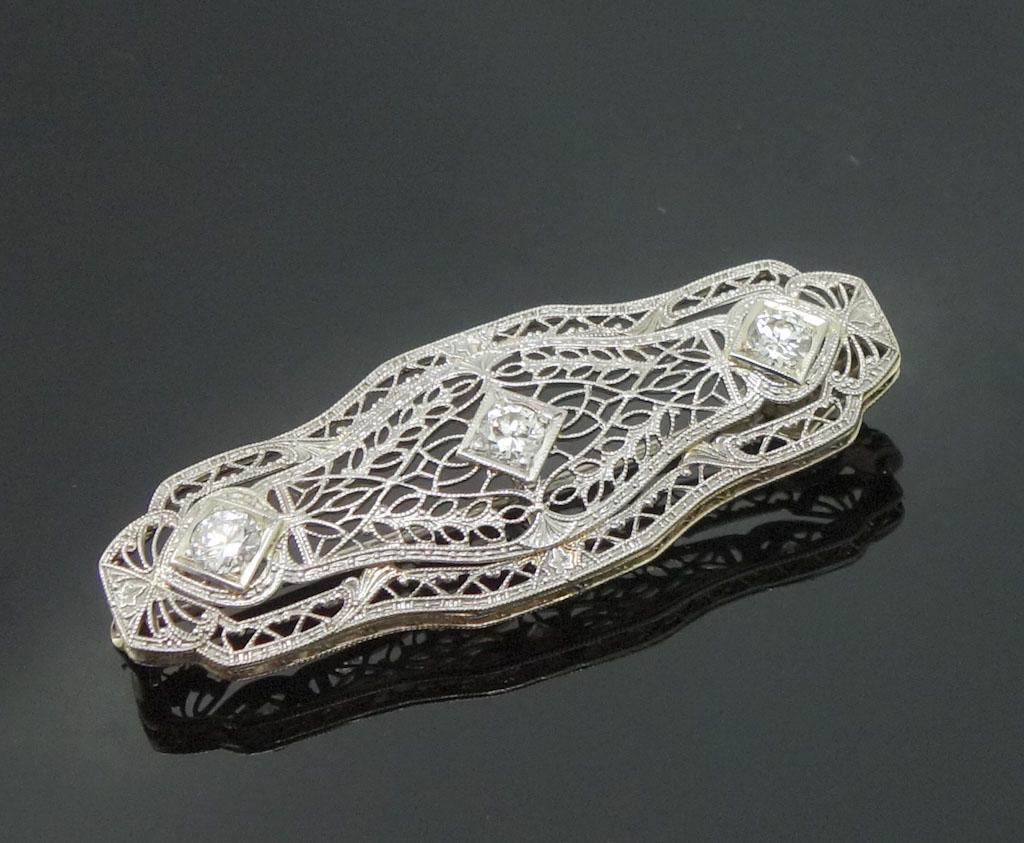 1920s 14k White Gold Filigree Diamond Brooch Pendant: This is an antique brooch pin/pendant dating to the 1920s. Rendered in fine 14k white gold, this Art Deco filigree brooch features a transitional cut diamond flanked by two Old European diamonds. A th