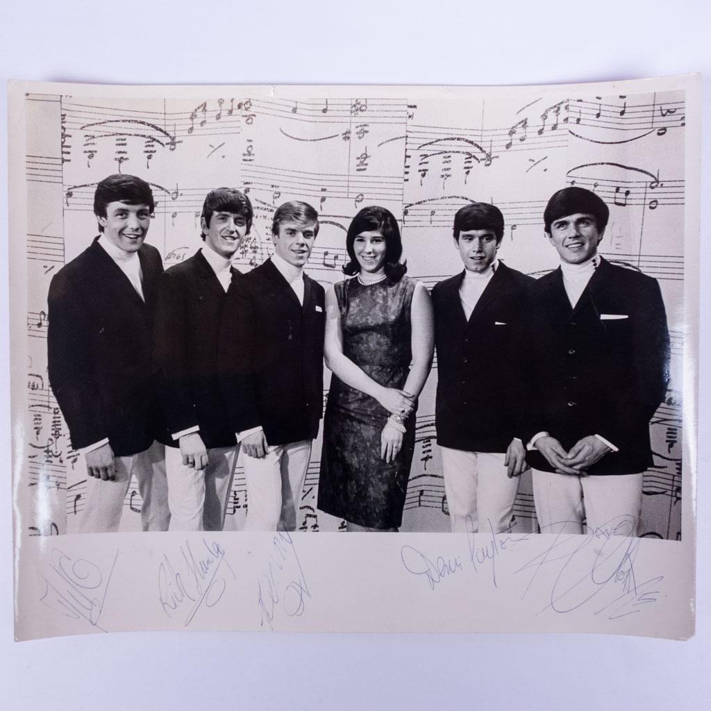 Autographed Signed Photo of Dave Clark Five Band: A photo of the band Dave Clark Five standing in a row with a fan in front of a music sheet backdrop. The photo is autographed by each member of the group. We guarantee the authenticity of all autograp
