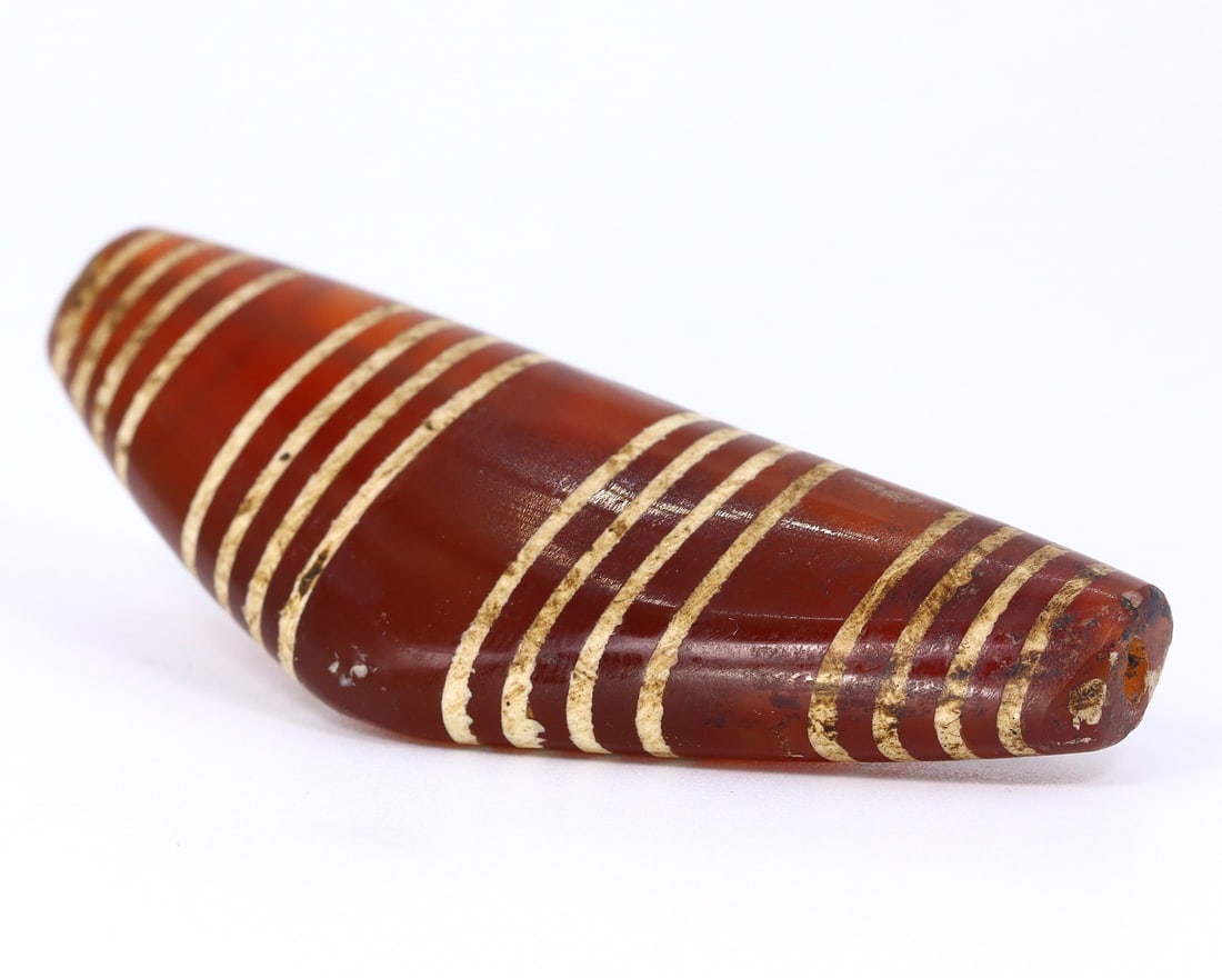 Antique Himalayan Carnelian Dzi Bead: Symbol of Protective Amulet (1650-1800AD): Dimension: 57.7x20.8mm Rarity Level: Very Rare Owing to its believed Tibetan origin and intricate stripe design, this dzi bead is not just an artifact but a relic that mirrors the deep spiritual and c
