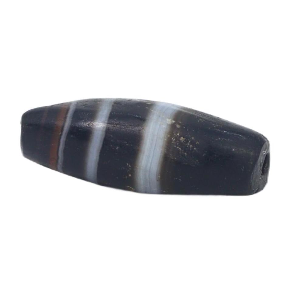 An Ancient Tibetan Banded Agate Chung Dzi Bead, 6th-10th Century AC: This chung dzi bead, with its enigmatic dark hues and stark banding, is a quintessential artifact of Tibetan culture. Dzi beads are deeply rooted in Tibetan tradition, often used as protective amulets