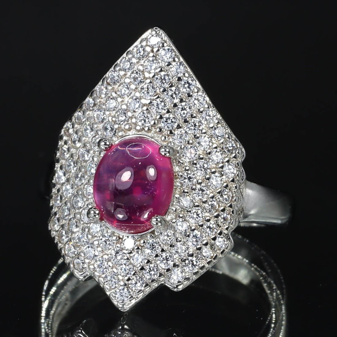 A Modern Silver Cocktail Ring with Natural Ruby, ca. 21st Century: This striking contemporary cocktail ring, stamped 925 silver, showcases a vibrant natural ruby set amidst a halo of sparkling cubic zirconia. Its bold design and bright gemstone make it a standout pie