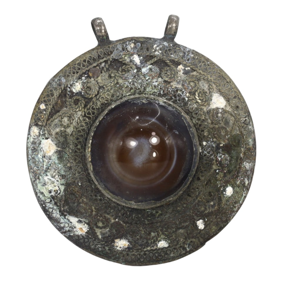 Himalayan Antique Circular Bronze Pendant with Luk Mik Goat Eye Stone: This circular bronze pendant features a central Luk Mik Goat Eye stone surrounded by intricate patterns, showcasing the ornate metalwork typical of Himalayan artisans. The symbolic inlay and detailed