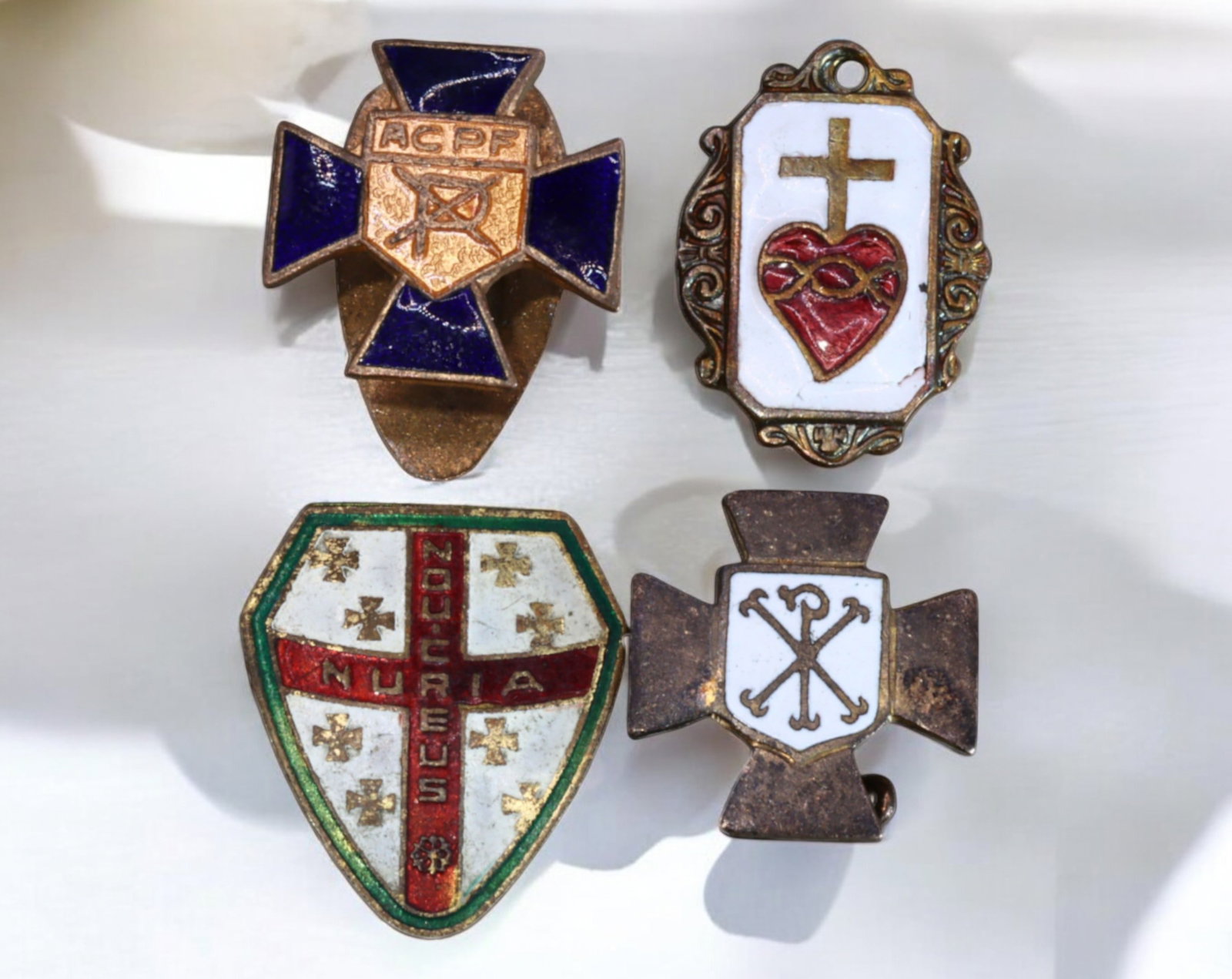SPAIN & FRANCE: Enameled Catholic Badge & Sacred Heart Medal Set (4 Pieces): A remarkable collection of four religious badges and devotional medals, reflecting European Catholic heritage. The set includes a shield-shaped enameled badge inscribed NURIA NOBILIBUS with a red cros