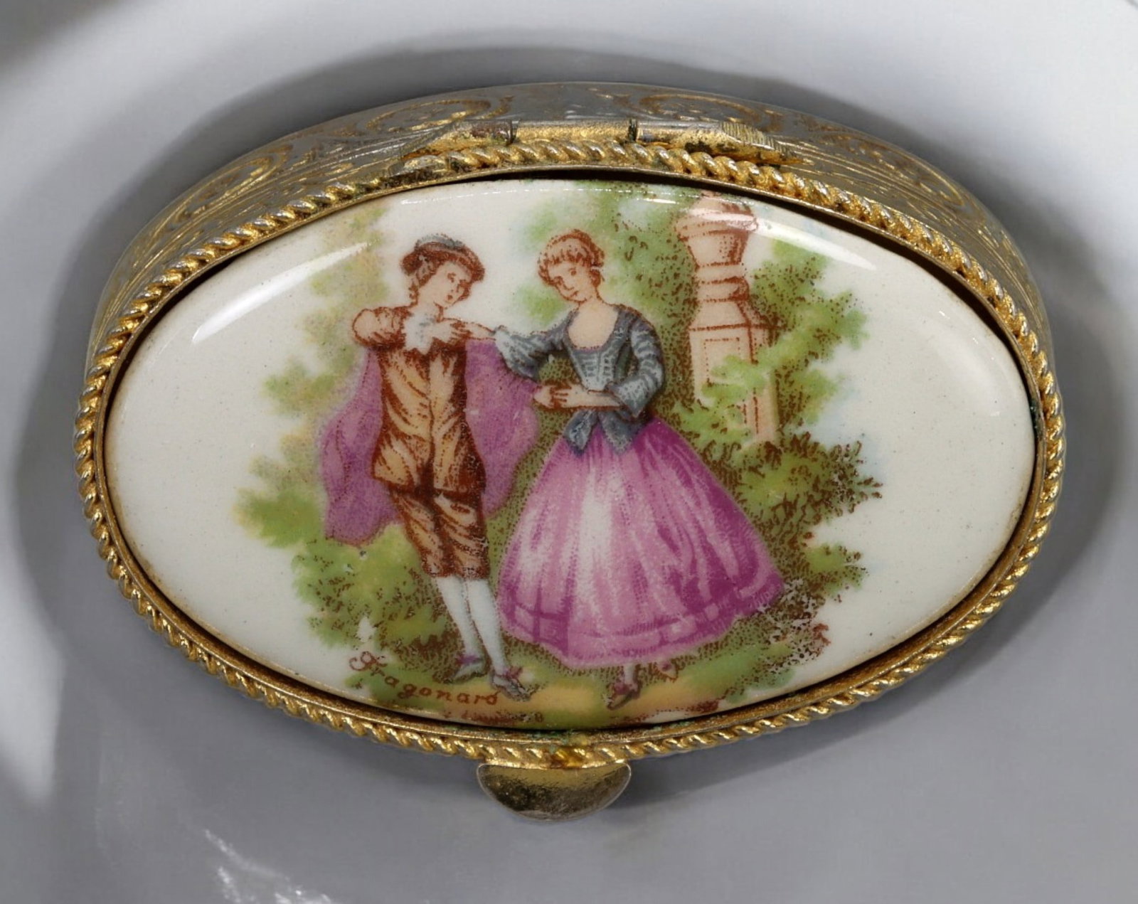 FRANCE: Fragonard-Style Porcelain Trinket Box - Gilded Metal Frame, 20th Century: A charming vintage trinket box, featuring a romantic Fragonard-style pastoral scene on a porcelain lid, encased in a gilded metal frame. Measuring approximately 5x1.5 cm and weighing 32g, this elegant