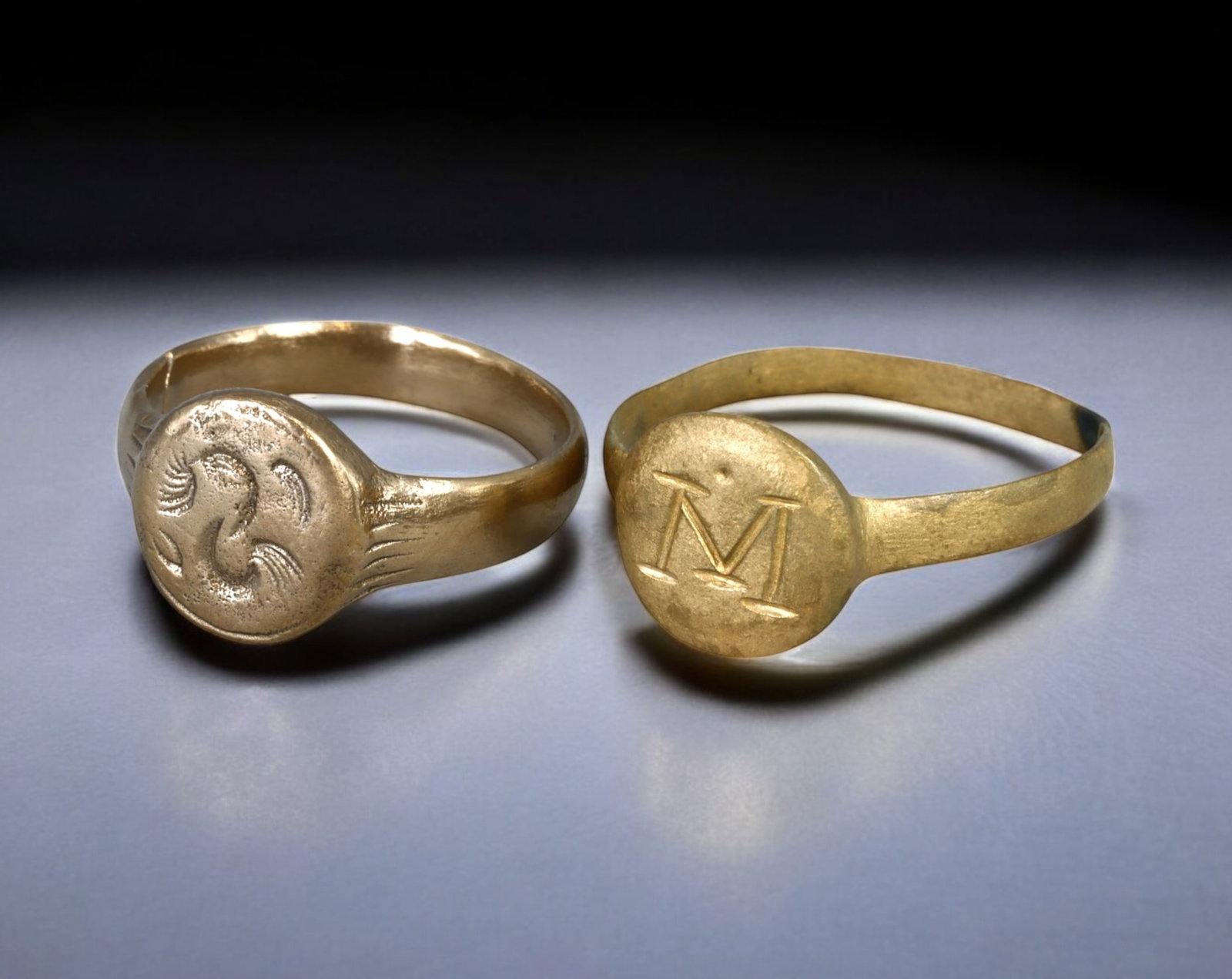 BYZANTINE: Pair of Bronze Signet Rings - 8.25-10 US, 7.7g, 6th-10th Century AD: An intriguing pair of Byzantine bronze signet rings, dating to the 6th-10th century AD, featuring distinct designs and historical significance. One ring bears an engraved "M" with a small central dot,