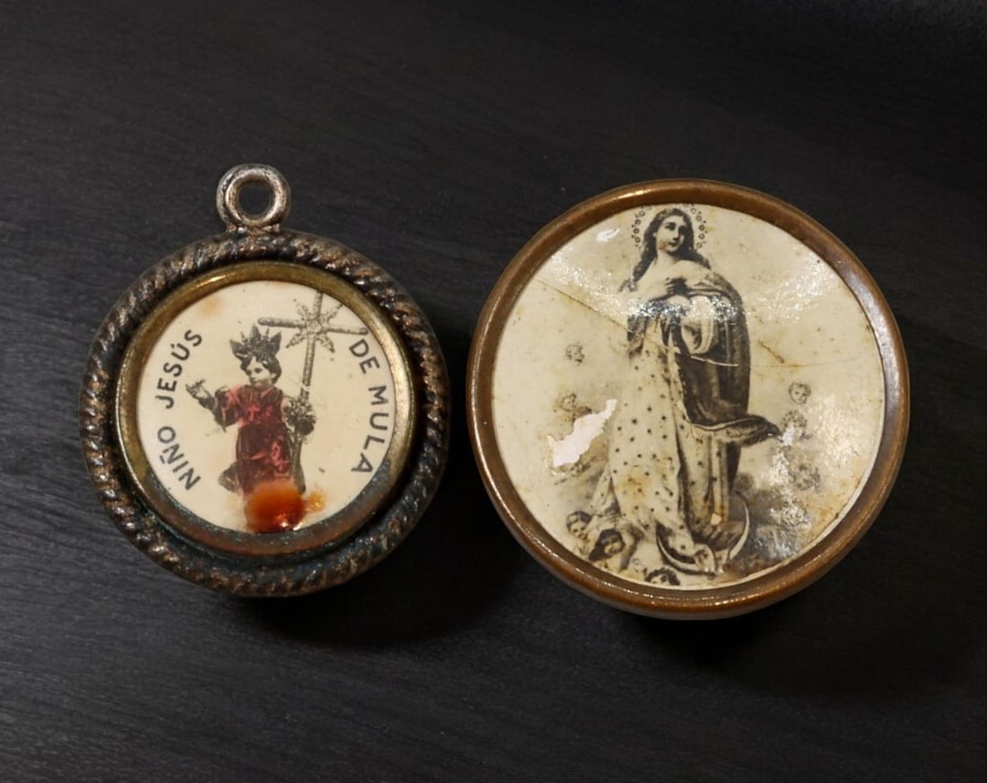 SPAIN: Nino Jesus de Mula & Virgin Mary Devotional Pendants - 19th Century: A pair of religious devotional pendants featuring printed images under glass, housed in metal frames. The first pendant showcases the revered Nino Jesus de Mula, a depiction of the Christ Child venera