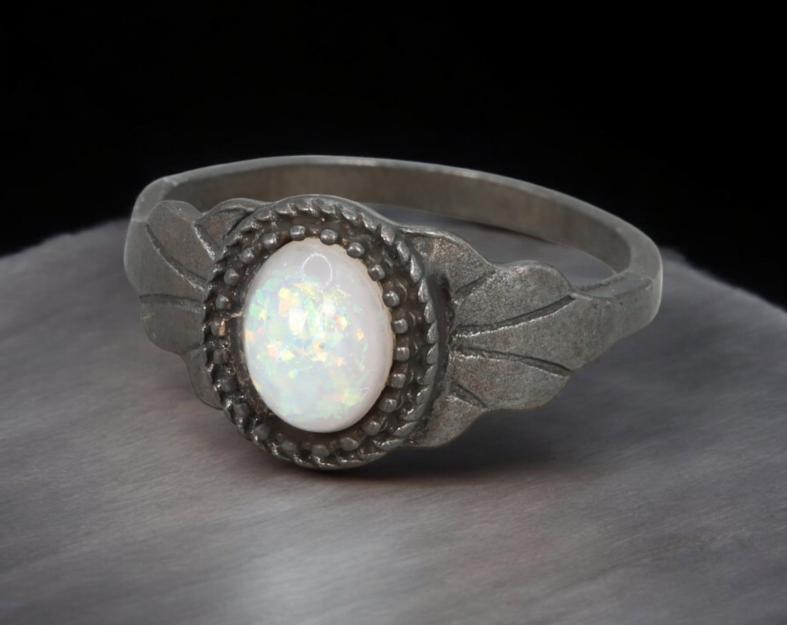 ARTS & CRAFTS MOVEMENT: Ring - Patinated Mount with Oval Iridescent Cabochon, US 8.7 (2.8g): A delicately handcrafted ring in the Arts & Crafts tradition, featuring a luminous oval cabochon with opalescent fire, framed by a beaded bezel and flanked with stylized leaf-like shoulders. The metal