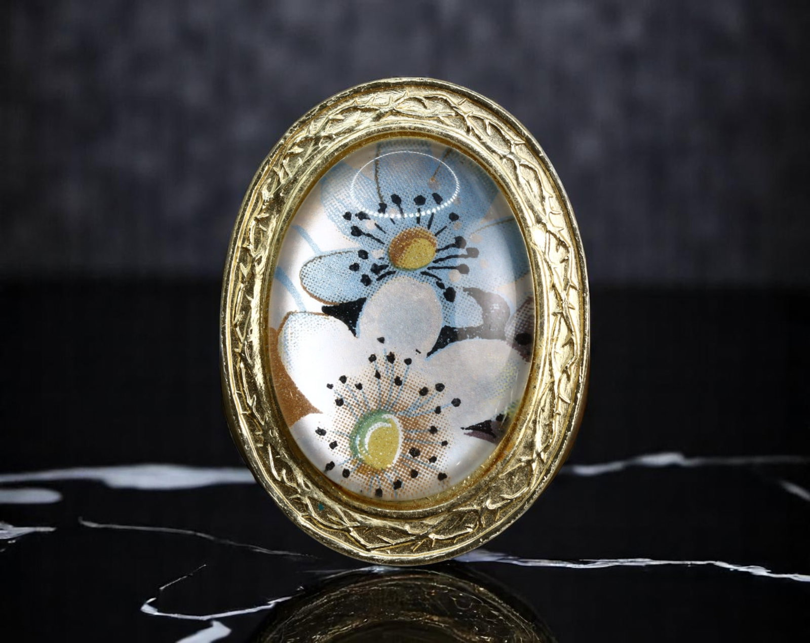 MID-CENTURY FLORAL OVAL BROOCH - Vintage Textile Under Glass, Engraved Goldtone Frame, 3.2x2.5cm: An evocative mid-century brooch featuring a stylized floral textile preserved beneath a domed clear covering, set in an engraved oval frame with bright goldtone finish. The fabric shows delicate bloss