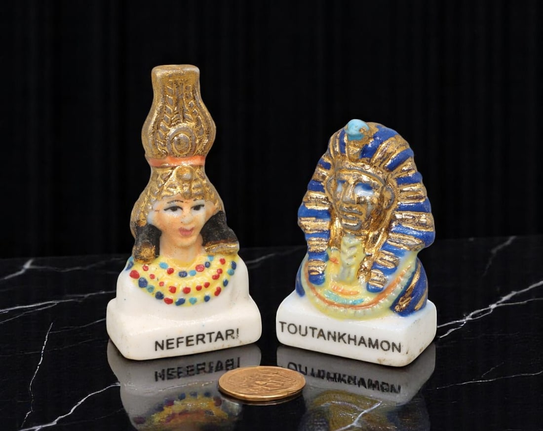 EGYPTIAN REVIVAL: Feve Pair - Nefertari & Tutankhamun, Gilded Porcelain by Banette (2 pcs, 2.5-3 cm): This visually arresting pair of hand-glazed feves celebrates ancient Egyptian royalty through the lens of 20th-century Egyptian Revival style, produced for the Banette bakery line. Each porcelain piec