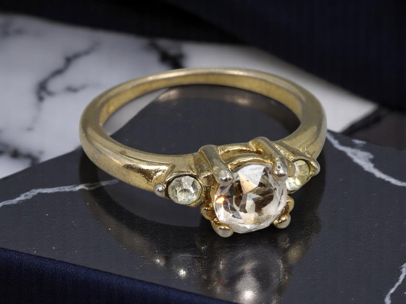 Three-Stone Ring - Clear Brilliant-Cut Stones, Stamped 18KT GE, Goldtone (Size 6.75 US): A classic contemporary three-stone ring featuring a luminous round brilliant-cut clear center stone flanked by two smaller clear accent stones. The smooth goldtone band is stamped "18KT GE," indicatin