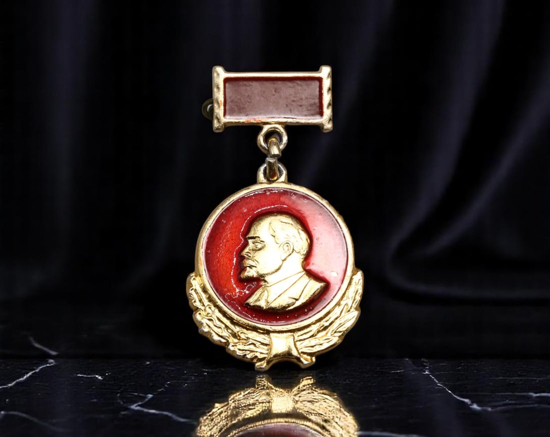 USSR: Honor of Labor - Lenin Award Badge with Red Enamel Suspension, ca. 1960s-1980s, 35x22mm: This distinctive Soviet award badge features a gold-tone portrait of Lenin set within a deep red enamel medallion, suspended from a matching rectangular red enamel bar. The laureate wreath motif at th