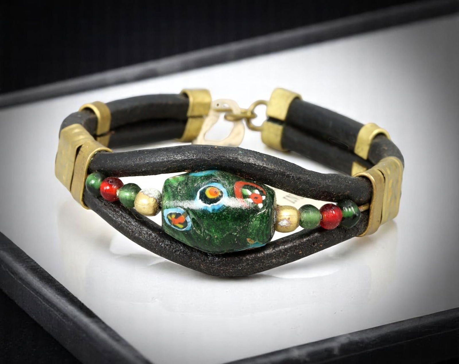 Tribal-Inspired Bracelet - Painted Beadwork & Leather Cords, Brass Accents: A richly textured mixed-media bracelet combining rustic black leather cords with a bold centerpiece bead featuring hand-painted, multicolor 'eye' motifs. Accented by smaller green, red, and gold-tone
