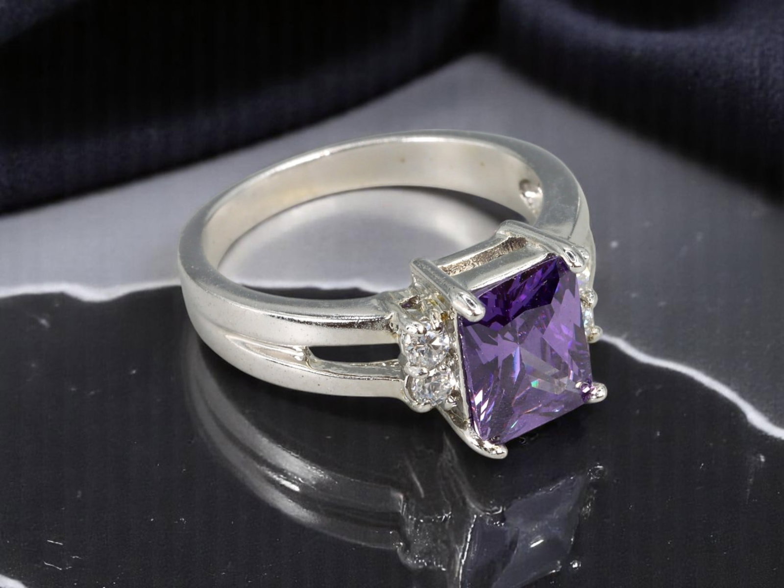 10KT Stamped Ring - Silvertone with Emerald-Cut Purple Center Stone & Clear Accents, US 7: A sleek and refined contemporary ring stamped 10 KT, featuring a bold emerald-cut purple center stone flanked by two sparkling clear accent stones. The split-shoulder band design and bright silvertone