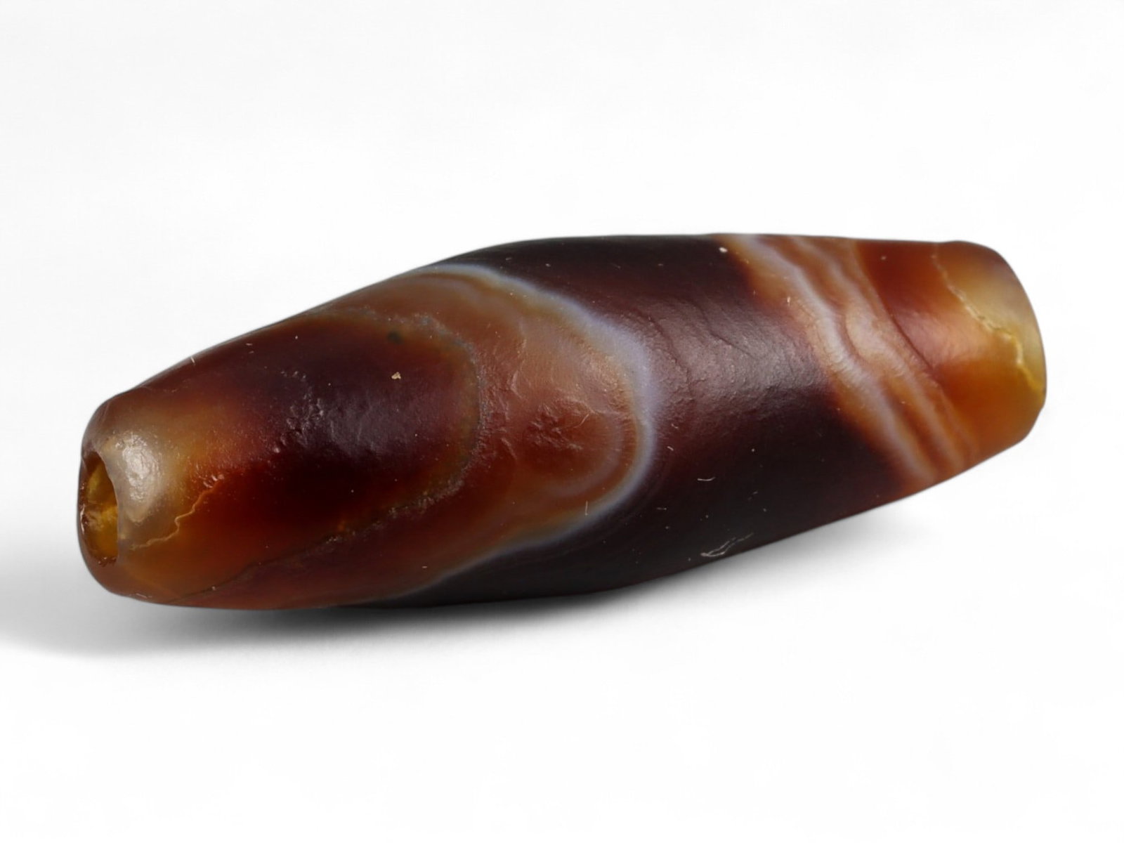 Ancient Tibetan Himalayan Banded Agate Dzi Bead, c. 9th-14th Century CE, 2.5cm: This ancient Tibetan Dzi bead, is carved from richly banded agate, its surface revealing bold layers of deep red, amber, and ivory-white. Its elongated cylindrical form, tapering at the ends, embodies