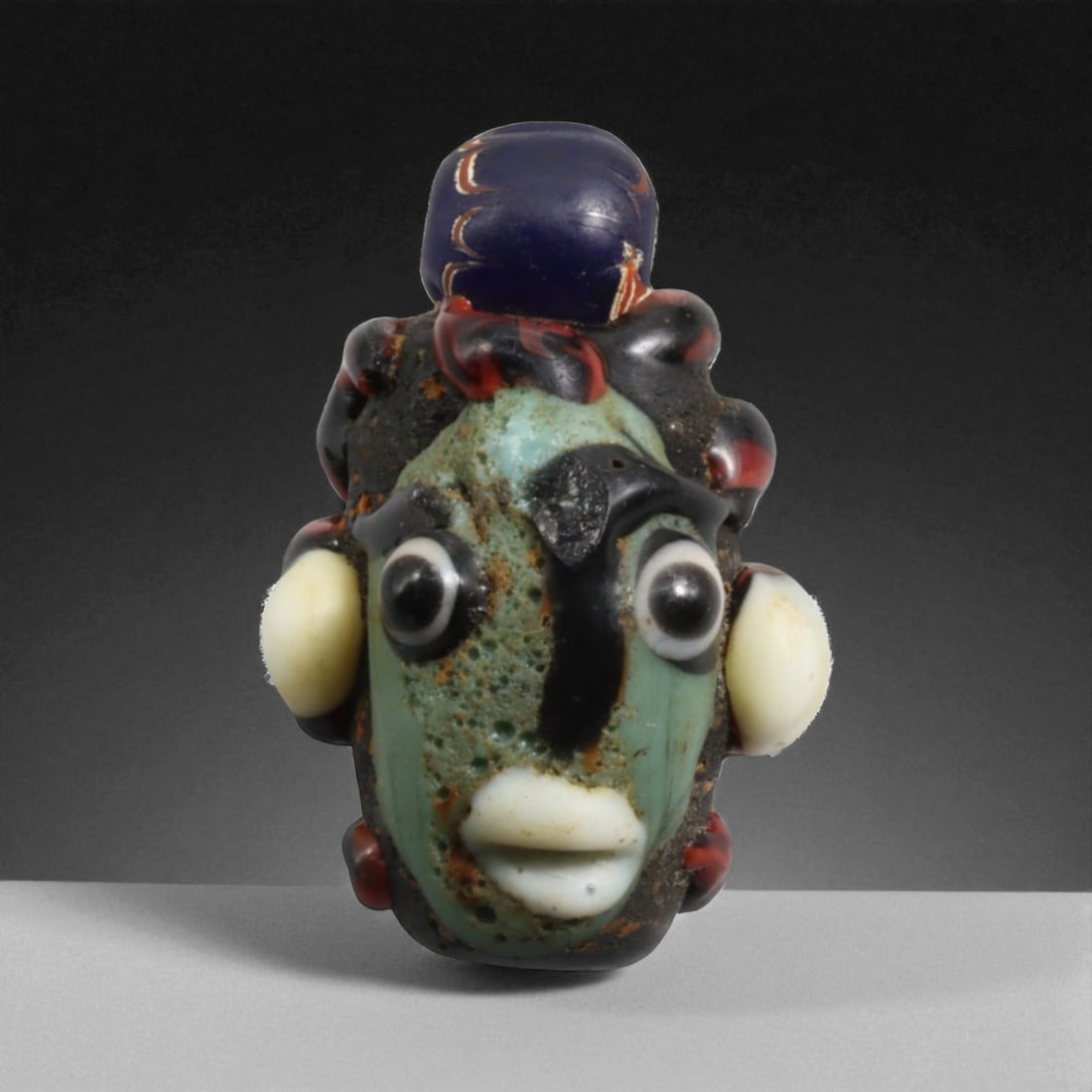 Ancient Phoenician-Etruscan Glass Head Pendant, c. 6th-4th Century BCE 12g: This remarkable glass head pendant bead, measuring 3 cm and weighing 12 grams, is a striking example of Phoenician and Etruscan cross-cultural artistry from the 6th-4th century BCE, with workshops in 