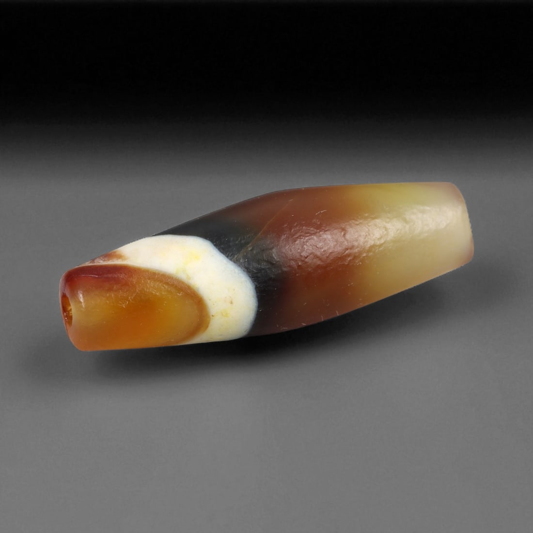 Ancient Tibetan Himalayan Banded Agate Dzi Bead, c. 9th-14th Century CE, 3.2cm: This ancient Tibetan Dzi bead, measuring 2.6 cm, is carved from richly banded agate, its surface revealing bold layers of deep red, amber, and ivory-white. Its elongated cylindrical form, tapering at 