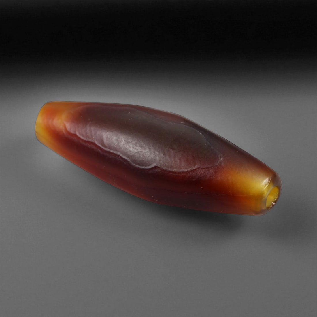 Ancient Tibetan Himalayan Banded Agate Dzi Bead, c. 9th-14th Century CE, 3.2cm: This ancient Tibetan Dzi bead, is carved from richly banded agate, its surface revealing bold layers of deep red, amber, and ivory-white. Its elongated cylindrical form, tapering at the ends, embodies