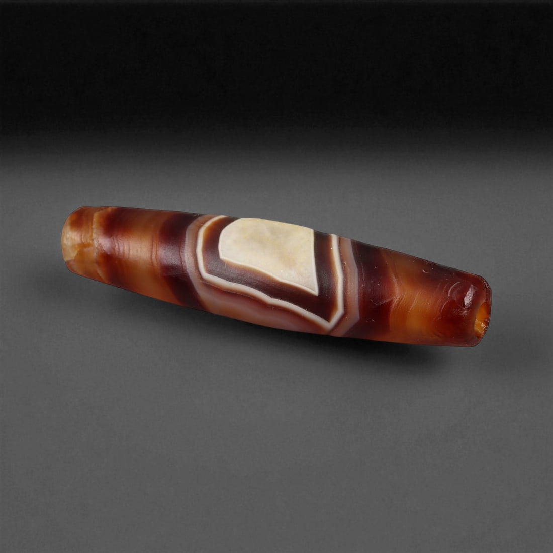 Ancient Tibetan Himalayan Banded Agate Dzi Bead, c. 9th-14th Century CE, 3.3cm: This ancient Tibetan Dzi bead, is carved from richly banded agate, its surface revealing bold layers of deep red, amber, and ivory-white. Its elongated cylindrical form, tapering at the ends, embodies