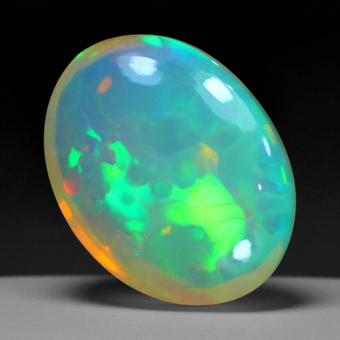 6.35 Carats Fiery Play-of-Color Welo Opal Oval Cabochon - Natural Ethiopian Stone: This mesmerizing Welo opal radiates a luminous play-of-color that dances across its translucent body in vivid flashes of red, orange, green, and violet 15.2x11.7x6.7mm. The gem's opalescent base glows