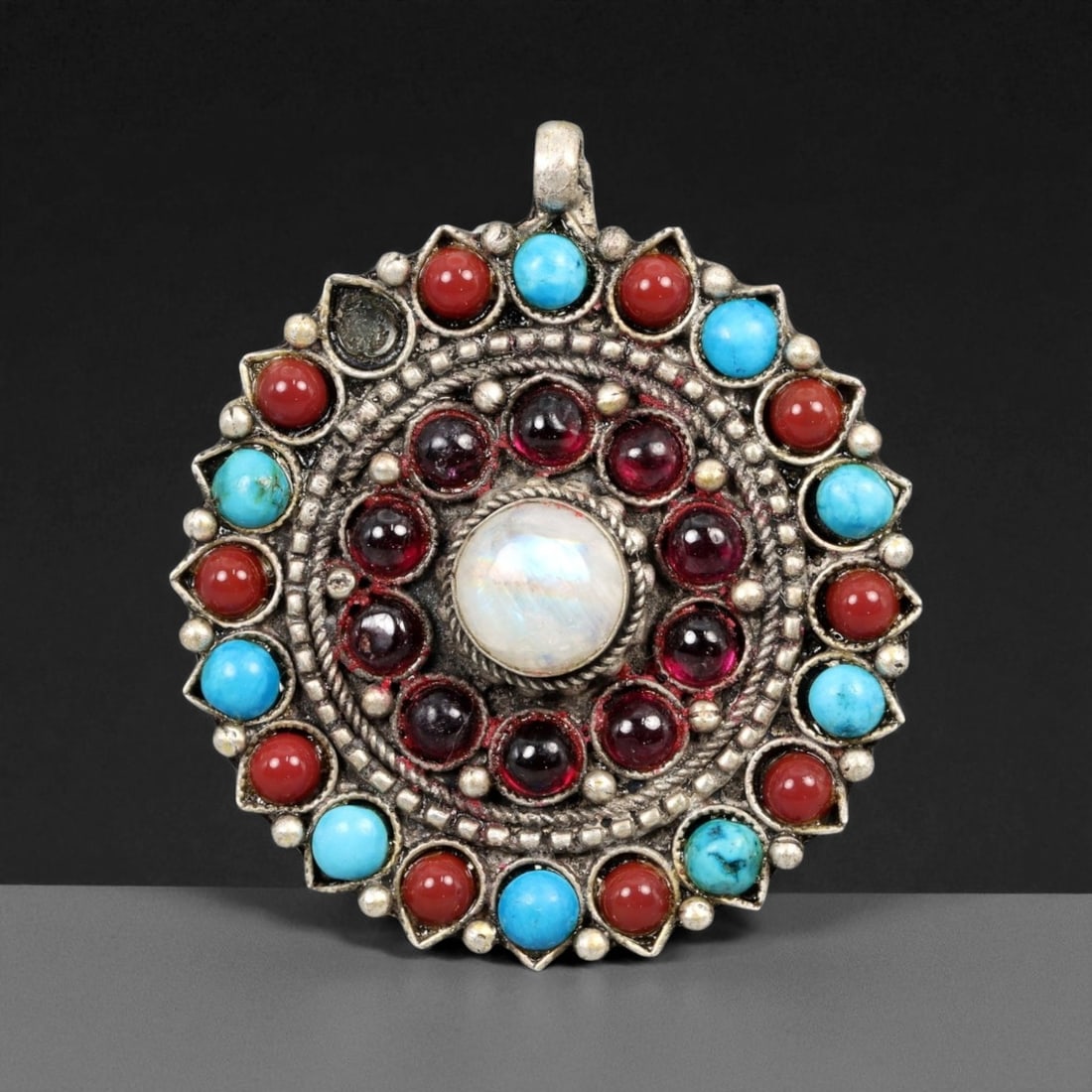 Vintage Himalayan Tibetan Moonstone Garnet Turquoise Coral Silver Pendant, 20th Century, 4.4cm: This vintage Himalayan Tibetan pendant radiates with vibrant color and symbolic depth, arranged in a circular mandala-like design. At its center rests a luminous moonstone cabochon, surrounded by a ri