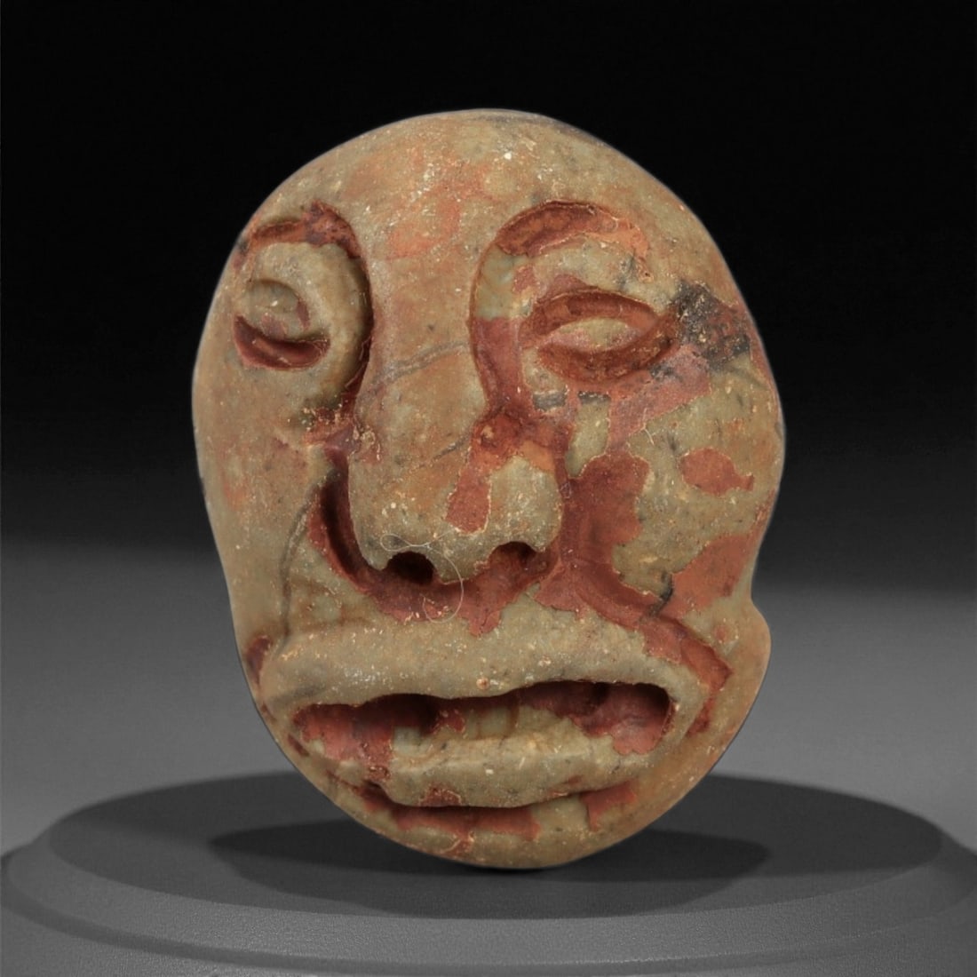 Ancient Medieval Coptic Egyptian Stone Maskette Amulet, c. 6th-9th Century CE, 2.6cm: This small but deeply evocative stone maskette, carved from pale brown stone with remnants of red pigment, dates to the Coptic Egyptian period of the 6th-9th centuries CE. The rounded facial form with