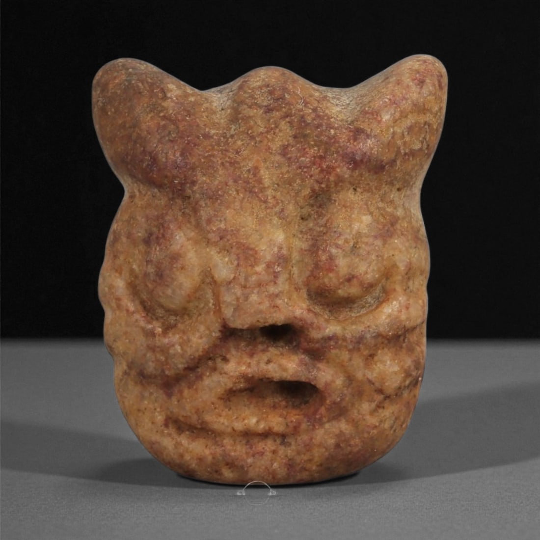 A Mediterranean Stone Maskette, 12th-17th Century, 19ZB: Carved from a mottled reddish-brown stone with granular surface texture, the maskette displays a compact, rounded form surmounted by three projecting points at the upper edge. The facial features are 