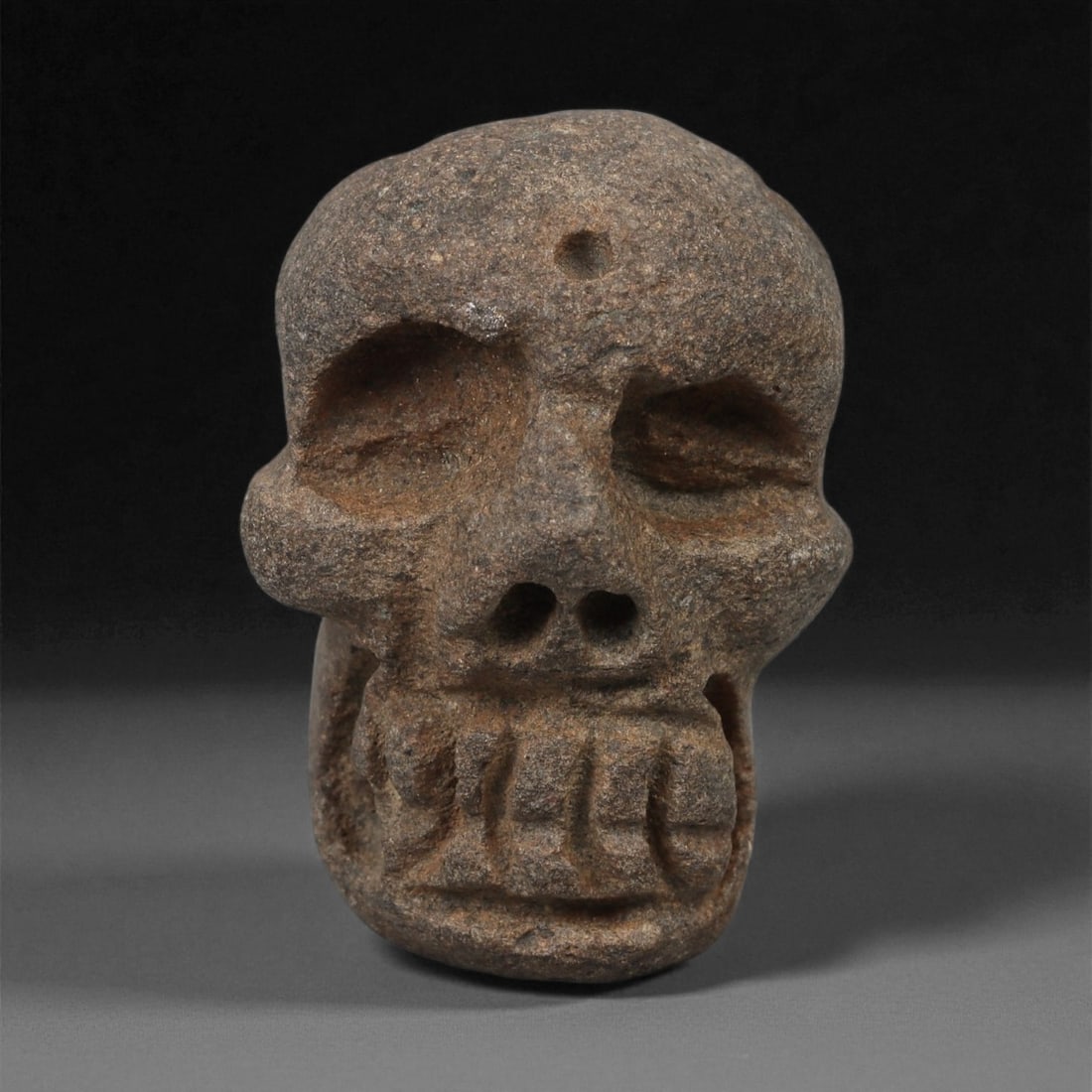 A Pre-Columbian Stone Skull Maskette, 1st Millennium CE, 78LA: Carved from dark brown stone with lighter surface inclusions, the maskette is shaped as a human skull with deeply recessed eye sockets and a defined nasal aperture formed by twin perforations. The mou