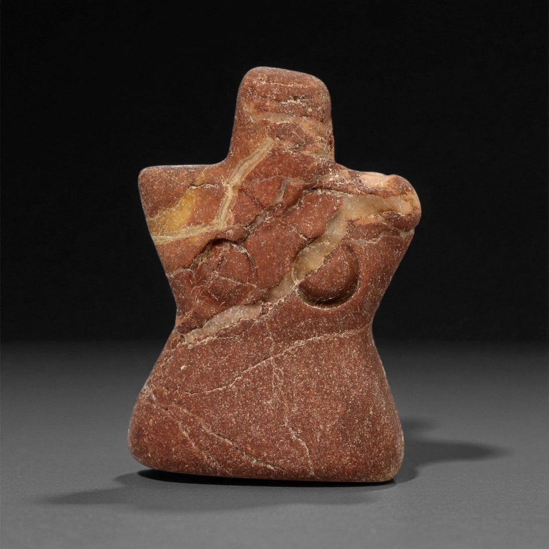 A Near Eastern Stone Anthropomorphic Figure, 3rd Millennium BCE, 83LA: Carved from reddish stone with lighter mineral veining, the figure is shaped with a broad lower body and a narrower upper section tapering to a short neck. The shoulders extend laterally to form angul