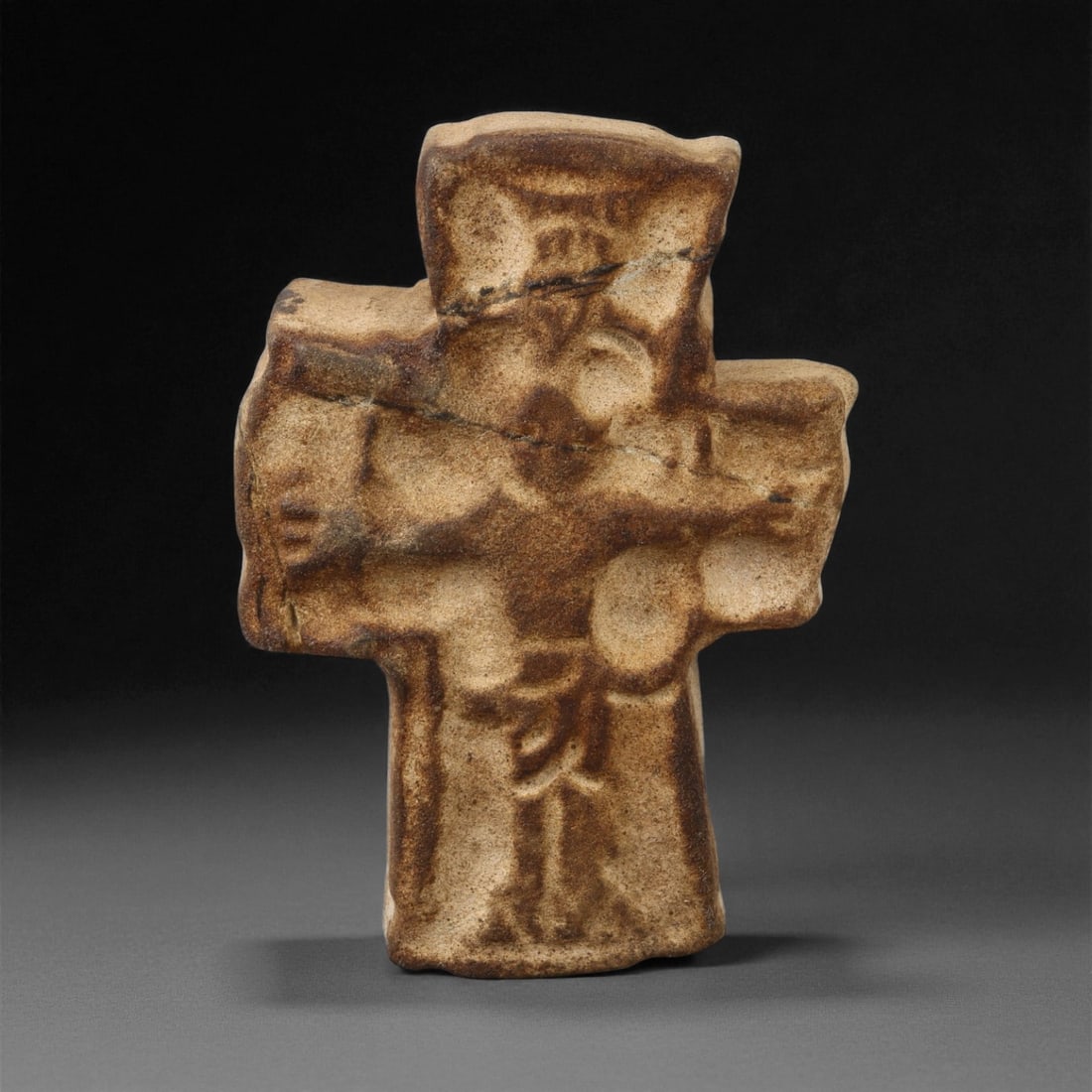 A Byzantine Stone Relief Cross with Figural Decoration, 6th-8th Century CE, 11LA: Carved from light brown stone with darker surface toning, the object is shaped as a cross with slightly flared arms. The front is worked in low relief with a central standing figure below the transver