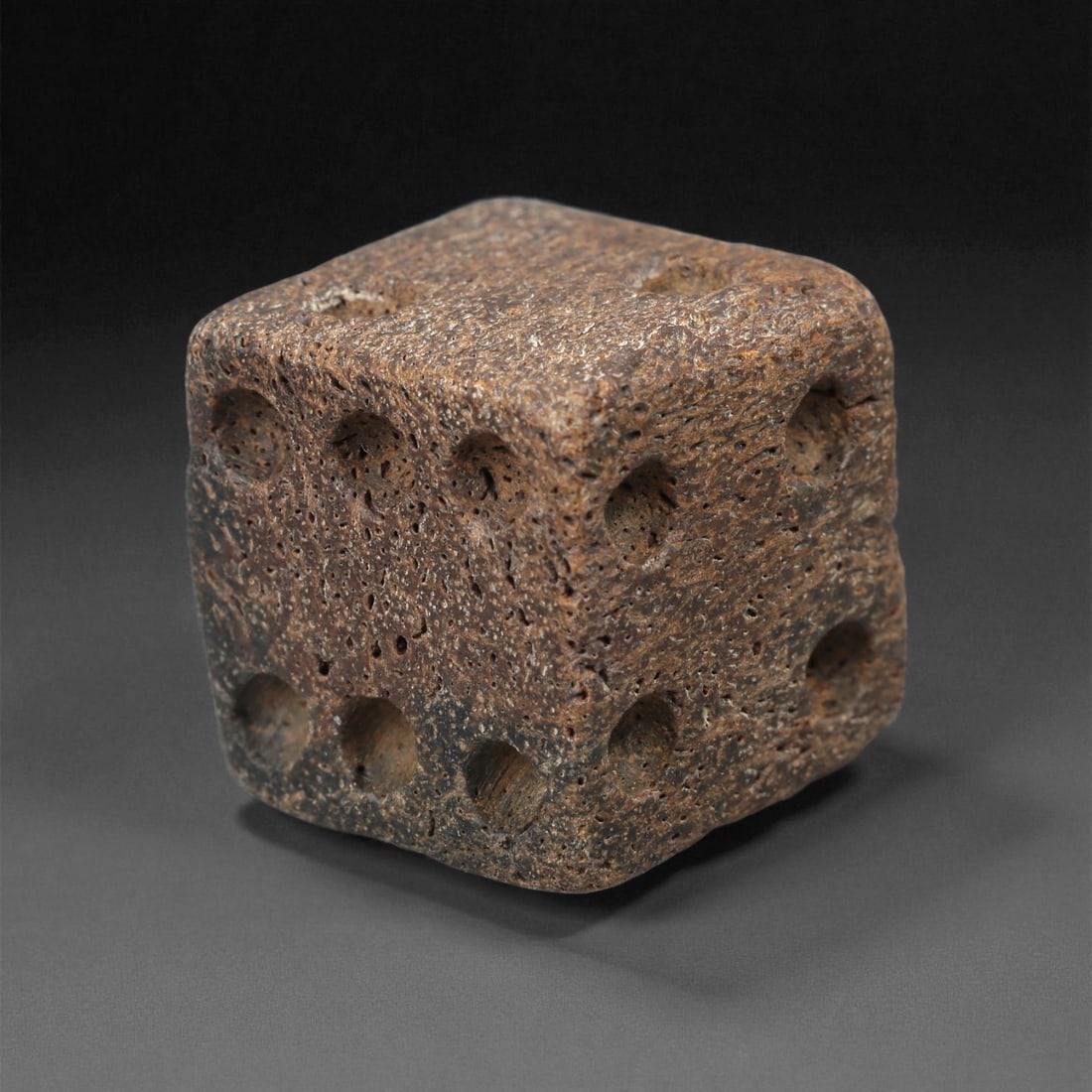 A Roman Bone Die, 1st-3rd Century CE, 5LD: Carved from light brown bone with a porous, naturally textured surface, the cubic die bears circular recessed pips on each face arranged in standard numerical configurations. The edges are slightly ro