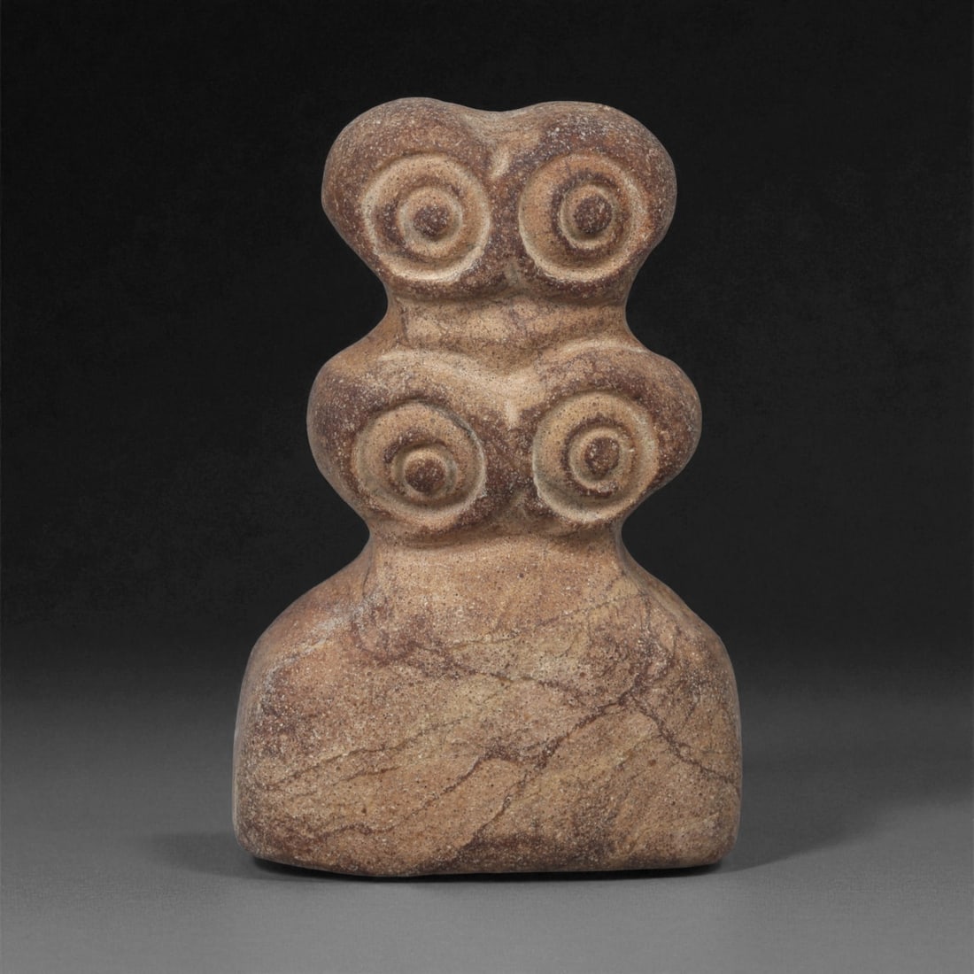 A Near Eastern Stone Eye Idol, 4th-3rd Millennium BCE, 22LA: Carved from light brown stone with natural veining, the figure is composed of a wide lower body surmounted by two stacked pairs of circular recessed eyes, each formed by concentric rings around a cent