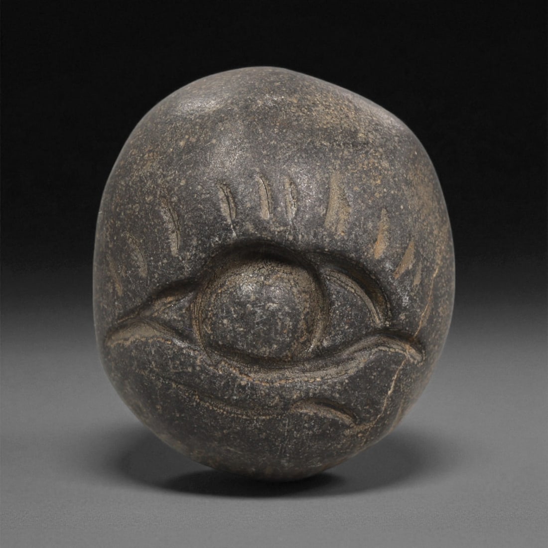 A Mediterranean Stone Maskette, 12th-17th Century, 96LC: Carved from a dark grey stone with lighter surface inclusions, the maskette is formed with a rounded contour. The design consists of a single large recessed eye rendered in pronounced relief, enclosed
