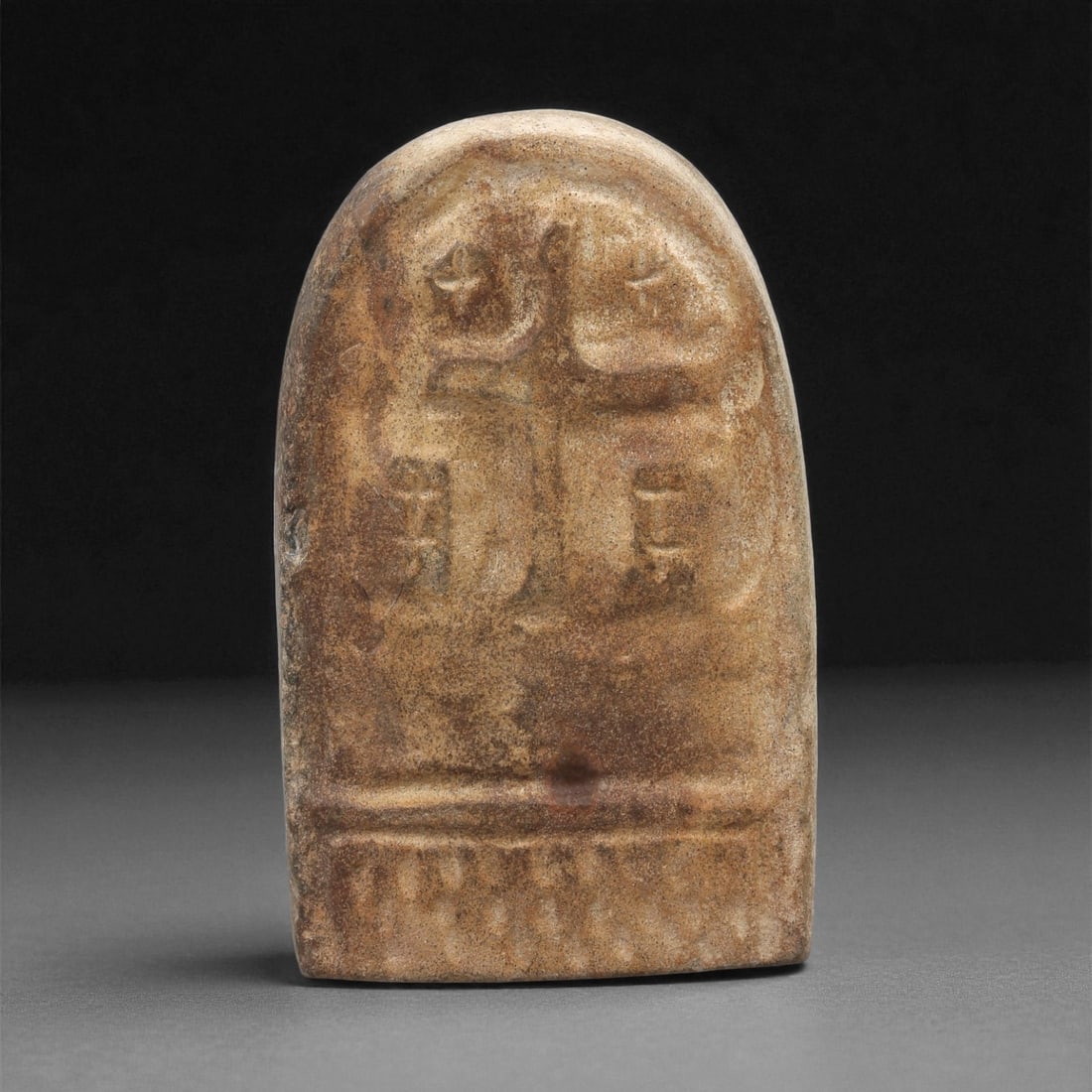 A Late Antique Stone Christian Stela, 4th-7th Century CE, 28LB: Carved from a light brown stone with natural mottling, the object is shaped as an upright stela with a rounded upper end. The front bears a low-relief Latin cross with slightly flaring arms, accompani