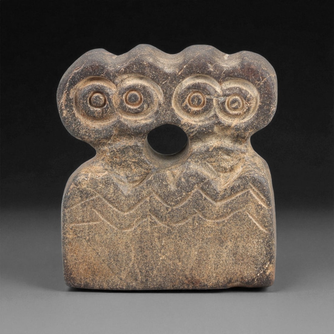 A Near Eastern Stone Eye Idol, 4th-3rd Millennium BCE, 19LD: Carved from a dark brown stone with lighter surface accretions, the idol features a rectangular lower body surmounted by a tri-lobed upper section bearing three pairs of recessed circular eyes. A dril