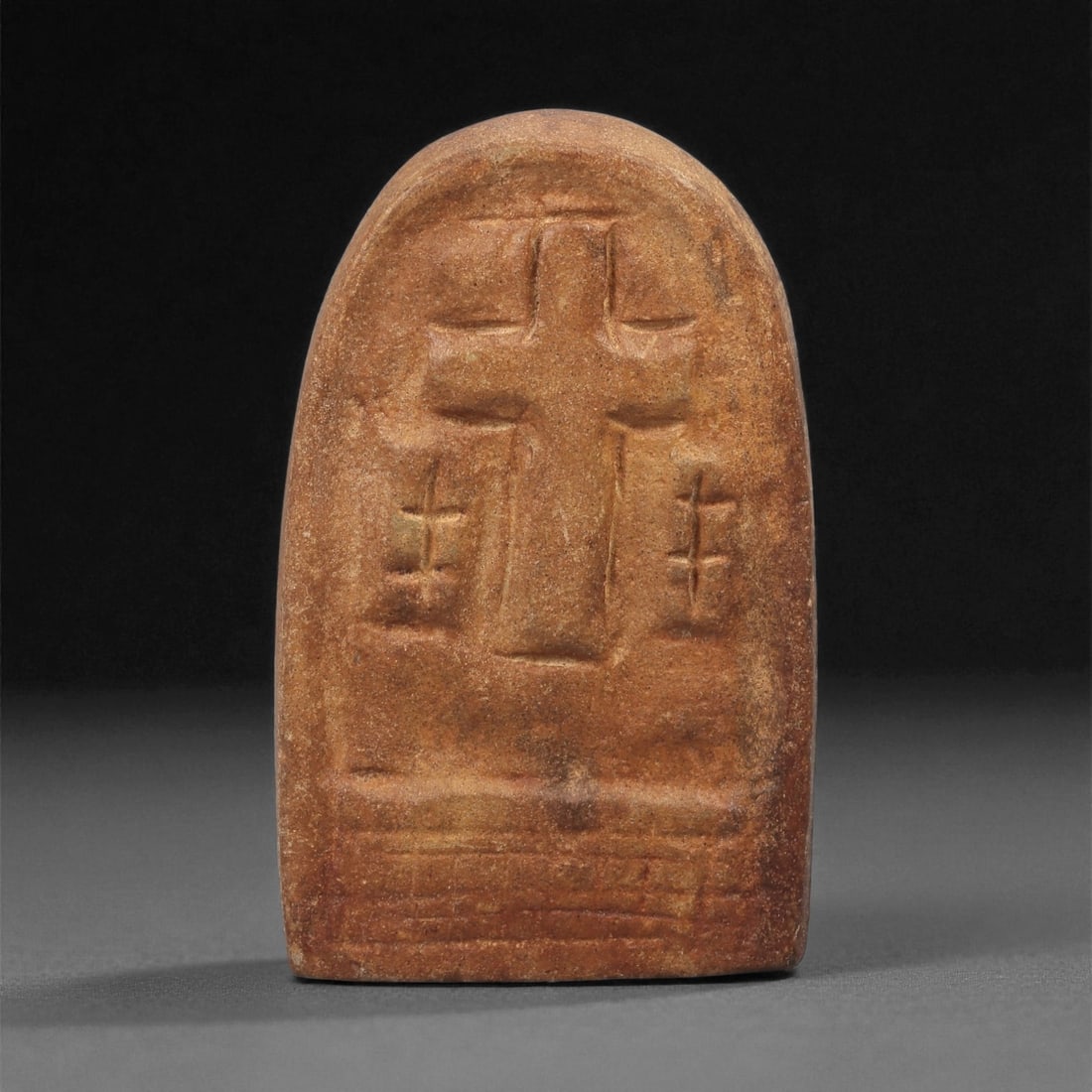 A Late Antique Stone Christian Stela, 4th-7th Century CE, 72LB: Carved from a reddish-brown stone with natural surface mottling, the object is shaped as an upright stela with a rounded upper end. The front bears a low-relief Latin cross with slightly flaring arms,