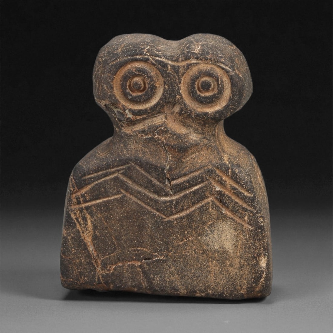 A Near Eastern Stone Eye Idol, 4th-3rd Millennium BCE, 72ZA: Carved from a dark brown stone with lighter veining, the figure is shaped with a rounded head bearing two recessed circular eyes formed by concentric grooves. A shallow depression is placed between th
