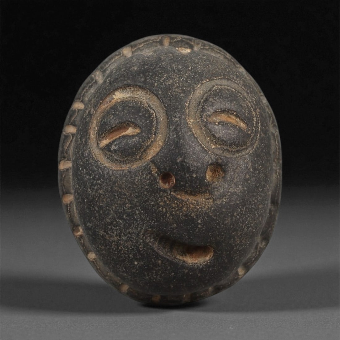 A Mediterranean Stone Maskette, 12th-17th Century, 39ZB: Carved from a dark grey stone with lighter surface abrasions, the maskette is formed with a rounded cranial contour and slightly tapered lower edge. The facial features are rendered in shallow relief,