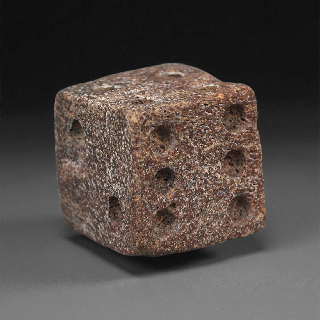 A Roman Bone Die, 1st-3rd Century CE, 6ZA: Carved from dense bone, the object is shaped as a small cube with slightly rounded edges. Each face bears a series of drilled circular pips arranged in standard numerical configurations. The surfaces 