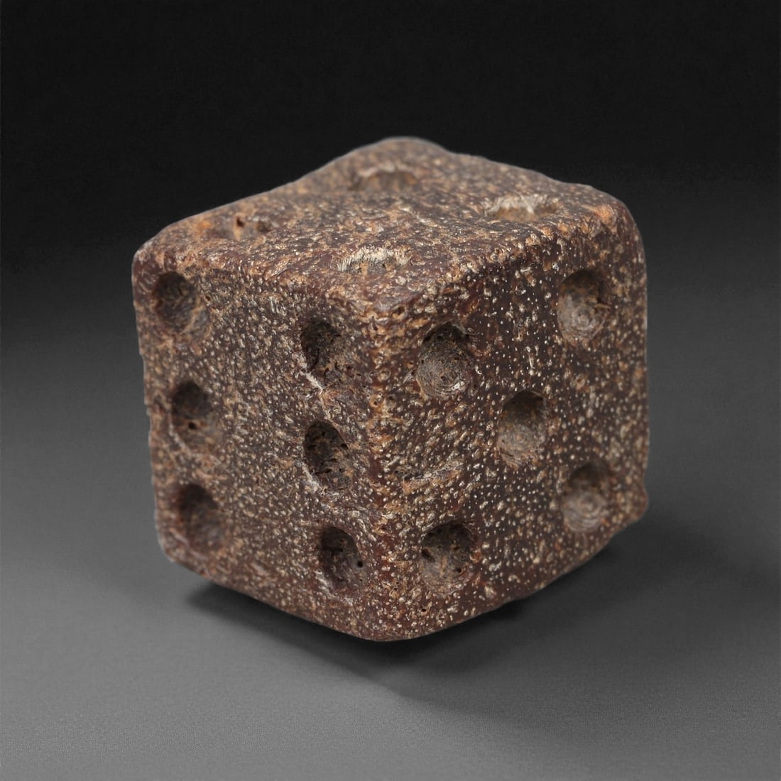 A Roman Bone Die, 1st-3rd Century CE, 11LD: Carved from light brown bone with a porous, naturally textured surface, the cubic die bears circular recessed pips on each face arranged in standard numerical configurations. The edges are slightly ro