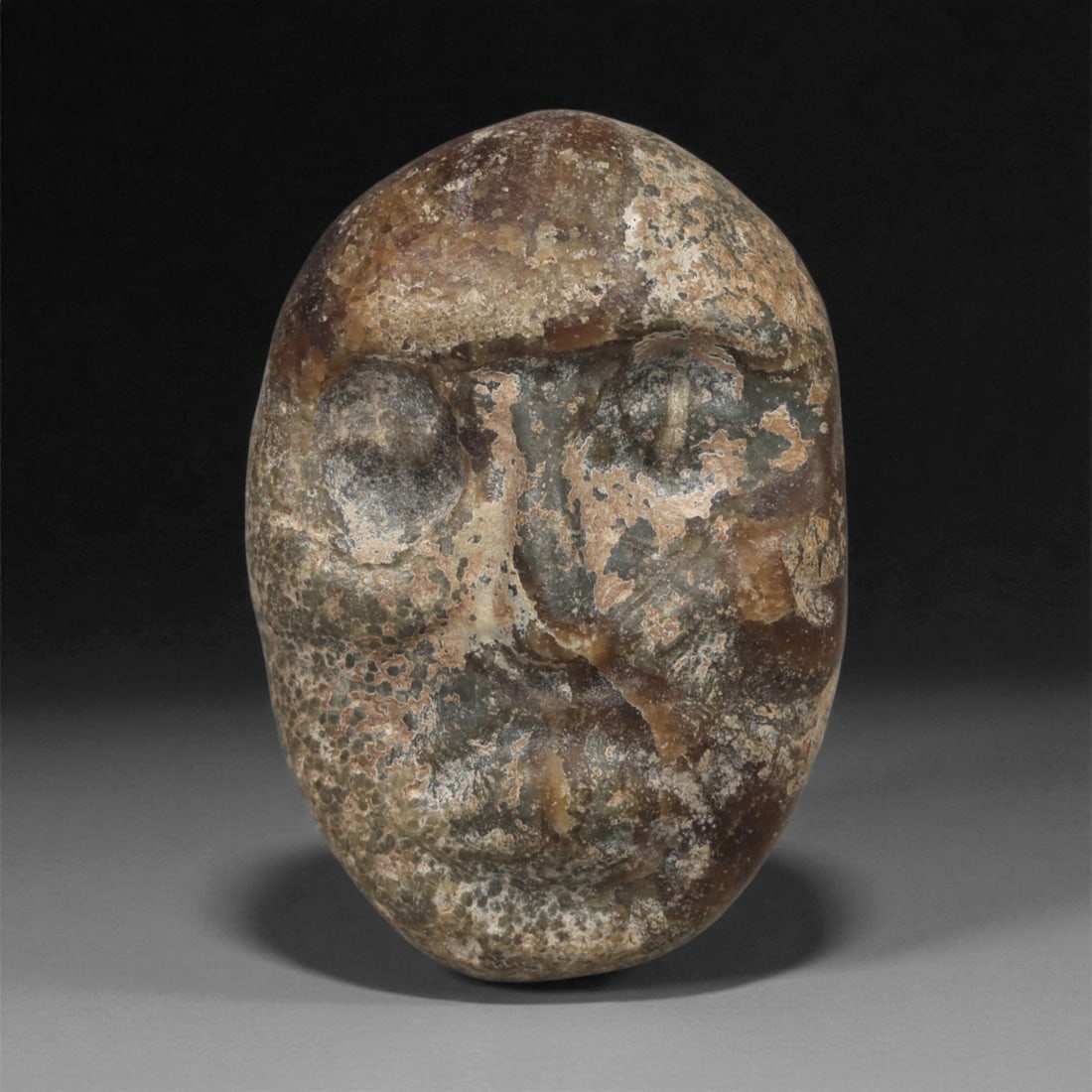 A Near Eastern Hardstone Maskette, 3rd-2nd Millennium BCE, 38LA: Carved from a mottled hardstone with areas of surface pitting and mineral accretion, the object is shaped as an oval maskette with a slightly convex front. The facial features are rendered in low reli