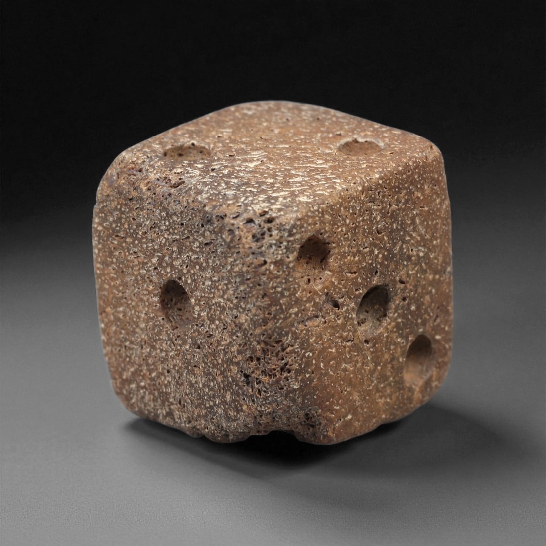 A Roman Bone Die, 1st-3rd Century CE, 36LD: Carved from light brown bone with a porous, naturally textured surface, the cubic die bears circular recessed pips on each face arranged in standard numerical configurations. The edges are slightly ro