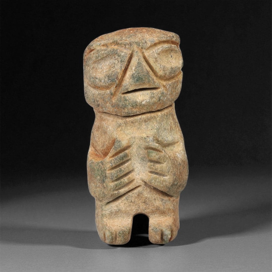 A Taino Carved Stone Idol, 1000-1500 CE, 39LE: Carved from light brown stone with areas of greenish surface accretion, the figure is modeled in a frontal stance with a compact body and short legs. The head is large in proportion, bearing deeply re