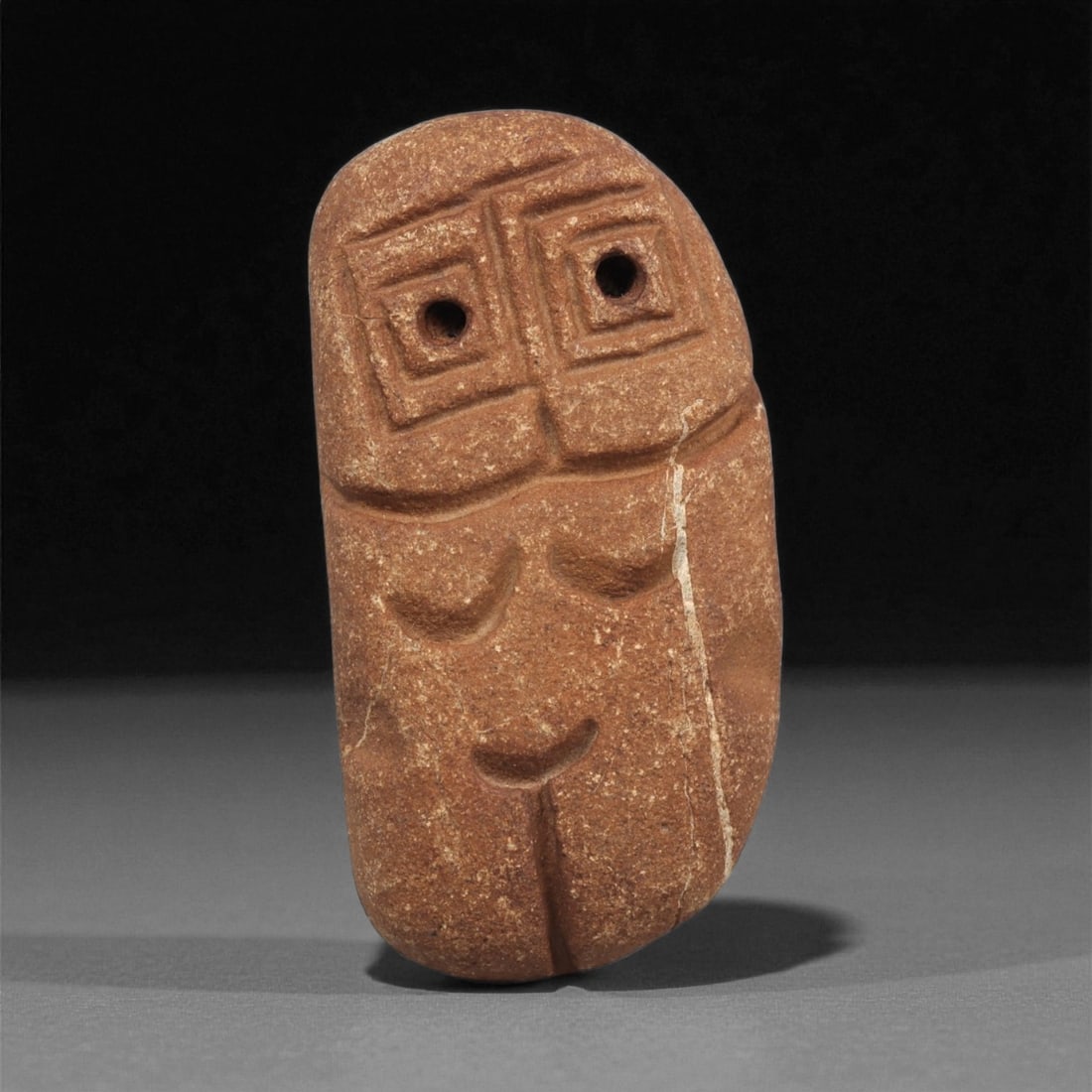 A Mediterranean Stone Maskette, 12th-17th Century, 8ZA: Carved from a reddish-brown stone with fine granular texture, the maskette is fashioned in a vertically elongated form with a flattened upper contour. The facial scheme is divided into two registers: 