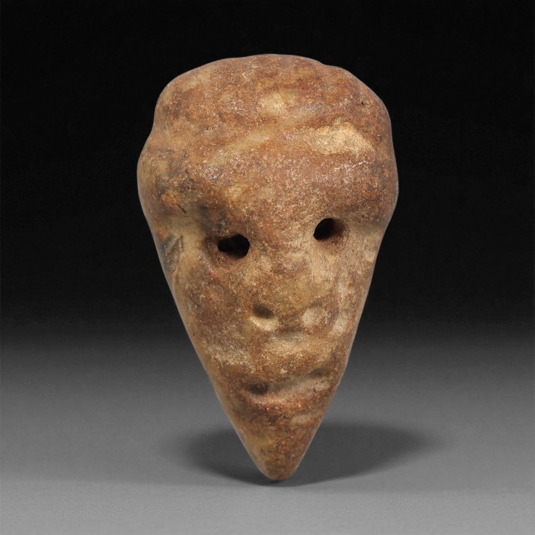 A Mediterranean Stone Maskette, 12th-17th Century, 64ZB: Carved from a light brown stone with natural surface variegation, the maskette is formed with a broad upper register tapering sharply to a pointed lower section. The facial features are minimally defi