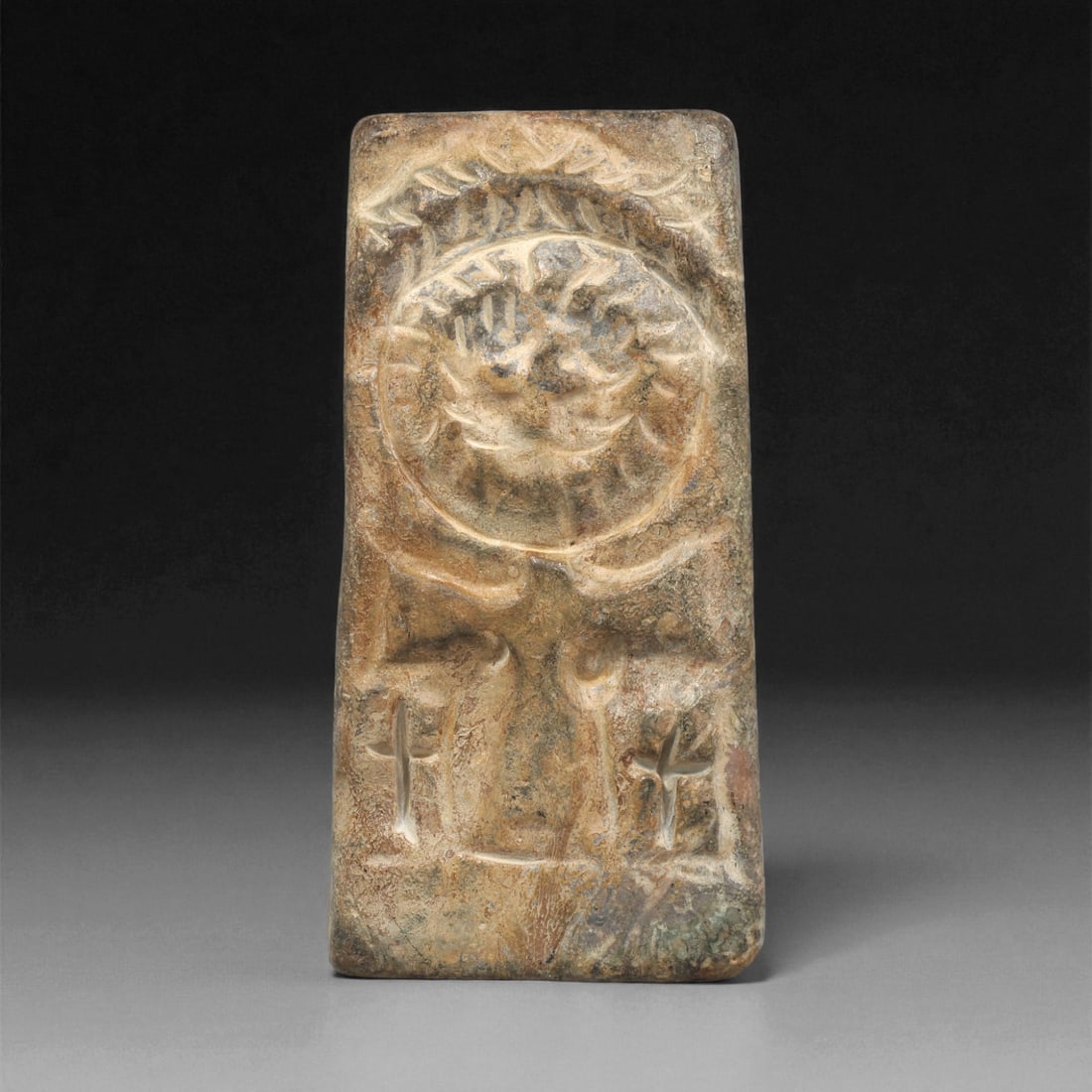 A Byzantine Stone Carved Plaque with Haloed Bust and Crosses, 6th-8th Century CE, 67LC: Carved from a light brown stone with surface patination and natural veining, the object is shaped as a tapering rectangular plaque. The upper register bears a shallow relief of a frontal haloed bust e