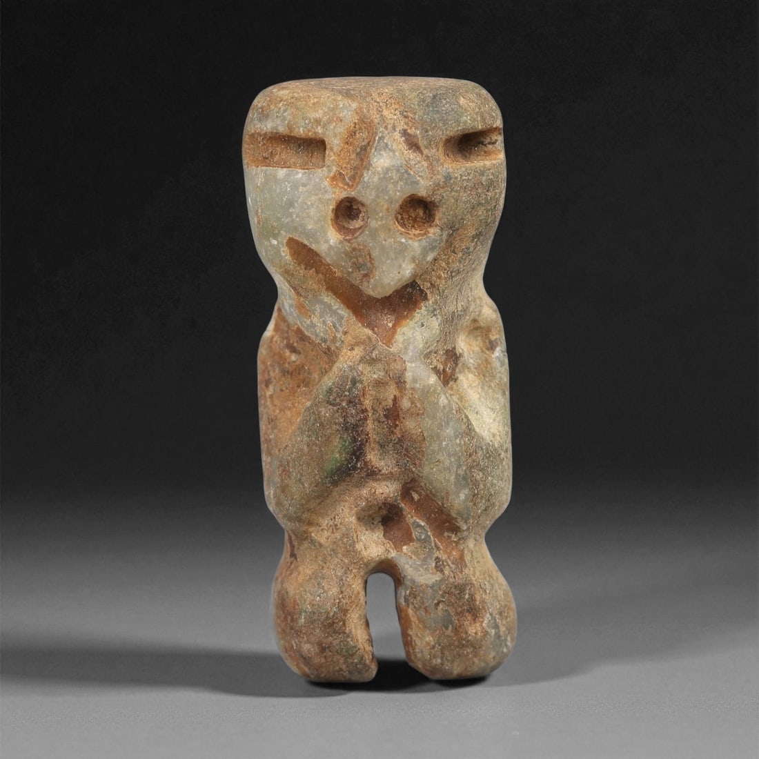 A Taino Carved Stone Idol, Late Ceramic Age, circa 1200-1500 CE, 28LD: Carved from mottled green-brown stone with surface weathering, the figure is shown standing with short bifurcated legs and a compact body. The head is rectangular with recessed horizontal eyes and dri